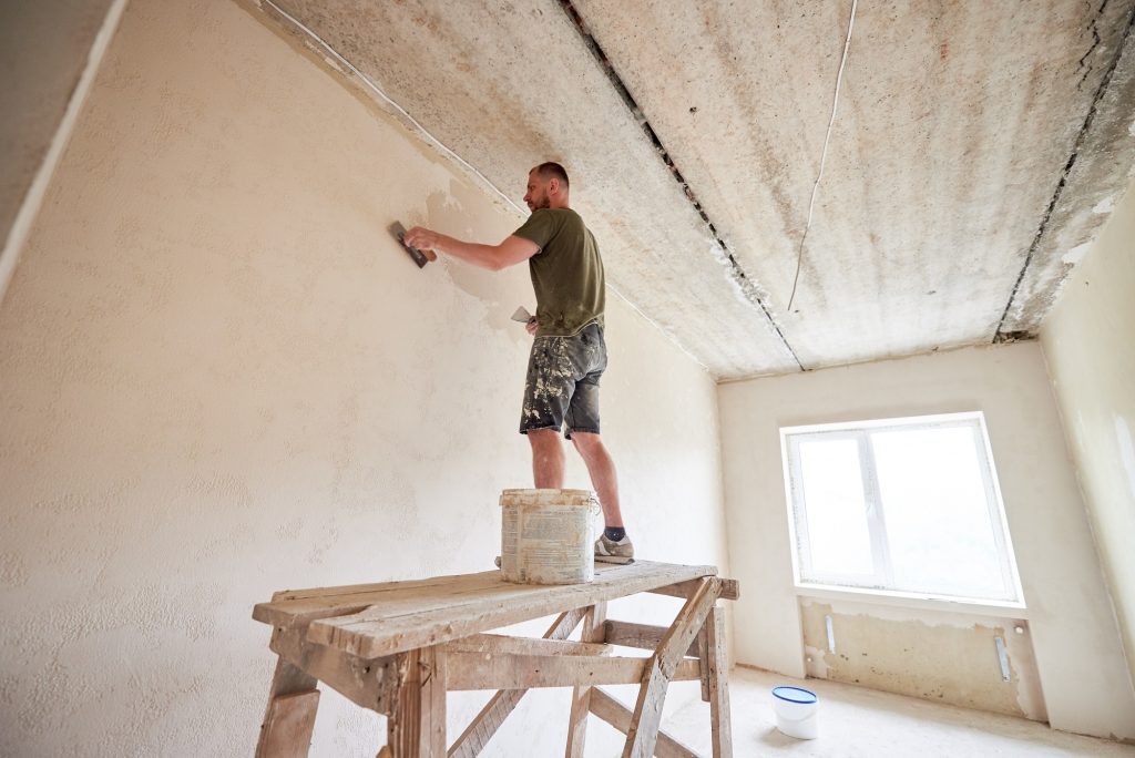 Young man is standing on wooden stand and working with spatula with plaster at the wall.