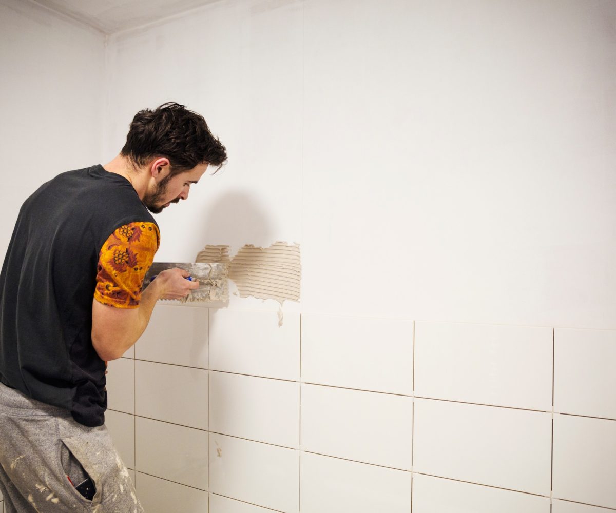 A builder, tiler placing white ceramic tiles on a wall in a bathroom.
